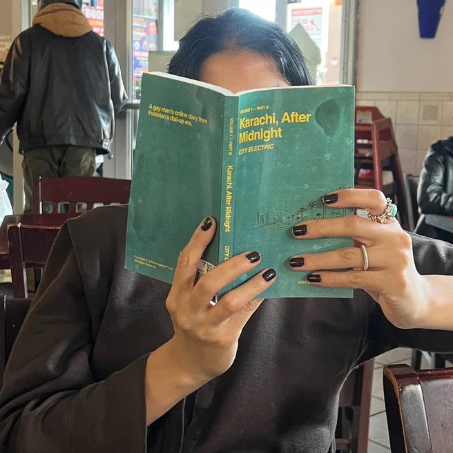 Person reading 'Karachi, After Midnight' book in a cafe, Khajistan archive theme