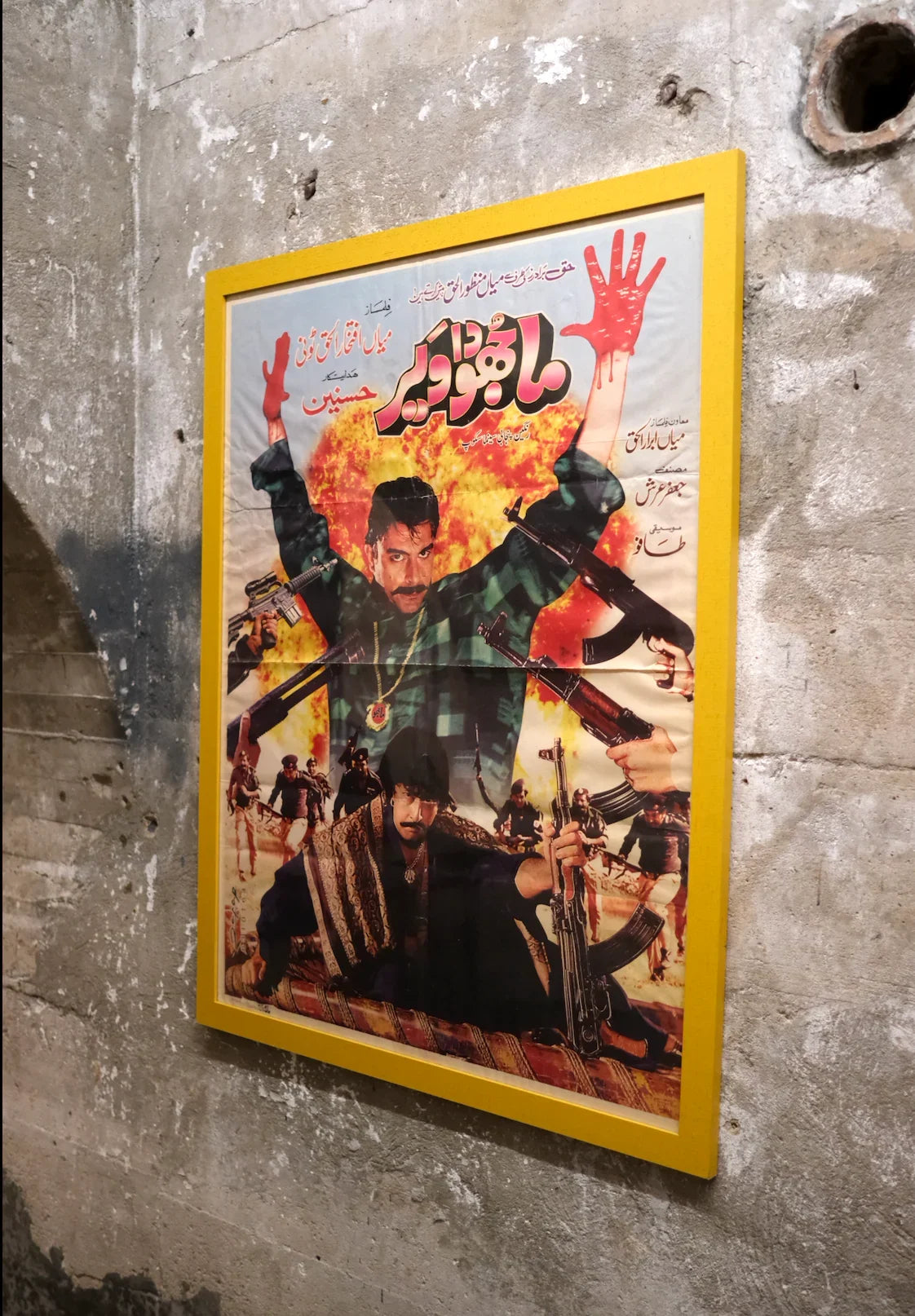 Vintage Pakistani movie poster with dramatic action scene and Urdu text, displayed on concrete wall at Khajistan.